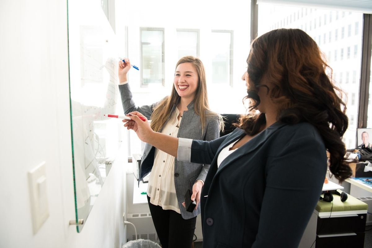 Two women writing and pointing at notes on a glass board in a bright office