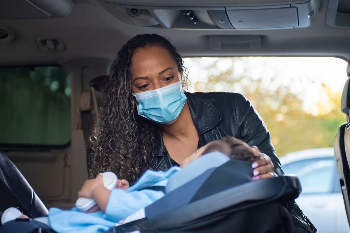 Parent in a face mask leaning over a baby secured in a black infant car seat inside a vehicle